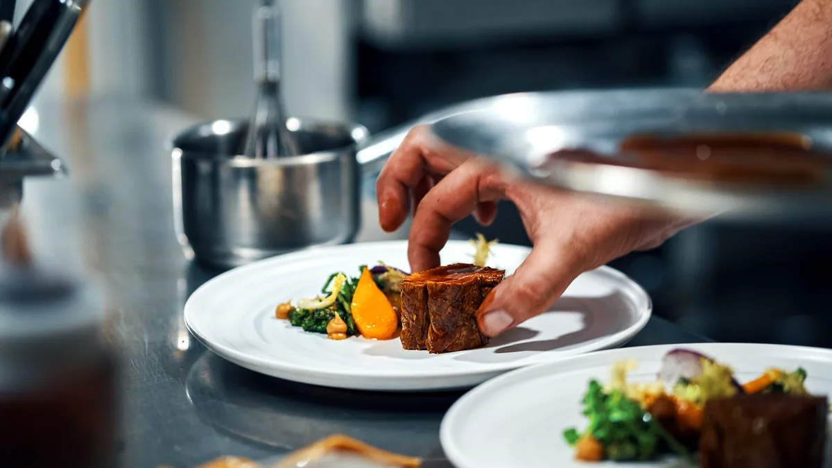 A chef's hands plating a dish in a busy commercial kitchen pass.
